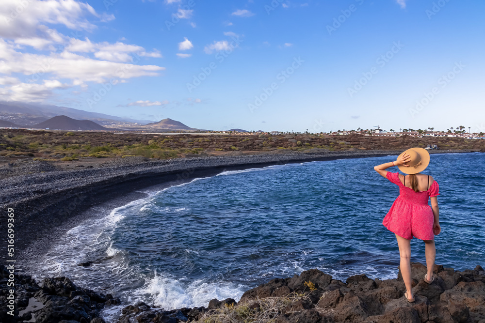 Woman with summer hat and red dress enjoying scenic view on black stone pebble beach Playa Colmenares near Amarilla, Golf del Sur, Tenerife, Canary Islands, Spain, Europe. Waves from Atlantic Ocean
