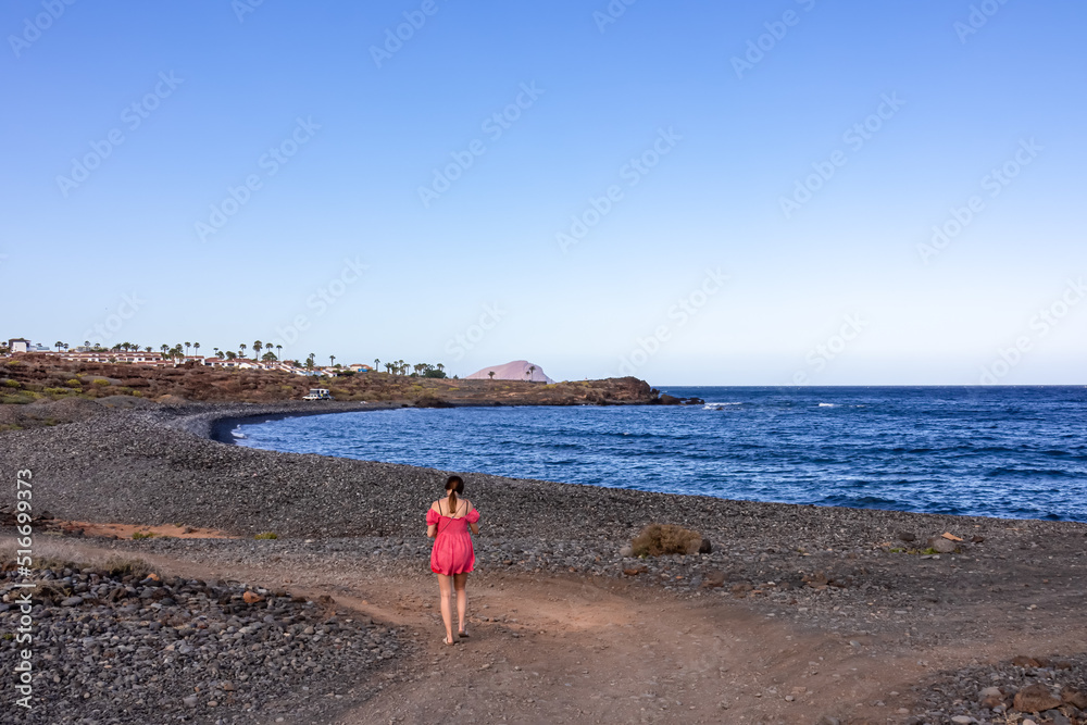 Woman in red dress walking on black stone pebble beach Playa Colmenares near Amarilla, Golf del Sur, Tenerife, Canary Islands, Spain, Europe. Waves from Atlantic Ocean. Scenic view on Montana Roja