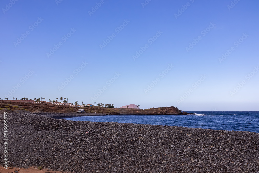 Scenic view on black stone pebble beach Playa Colmenares near Amarilla, Golf del Sur, Tenerife, Canary Islands, Spain, Europe. Vacation resort complex. Waves from Atlantic Ocean. Montana Roja in back Scenic view on black stone pebble beach Playa Colmenares near Amarilla, Golf del Sur, Tenerife, Canary Islands, Spain, Europe. Vacation resort complex. Waves from Atlantic Ocean. Montana Roja in back