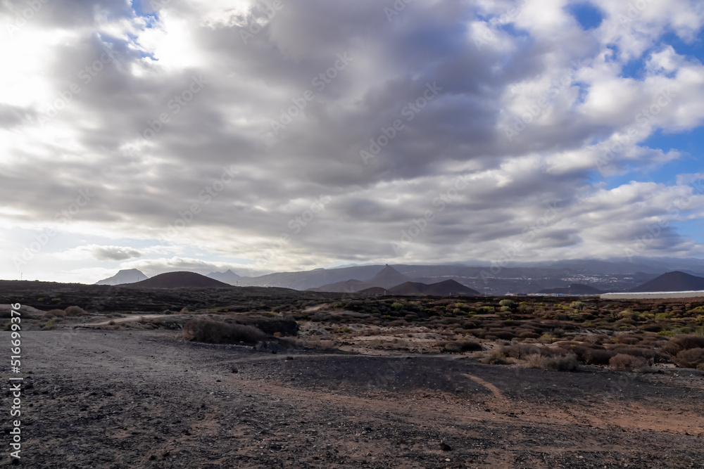 Panoramic view on cloud covered Pico del Teide and volcanic crater landscape seen from beach Playa Colmenares near Golf del Sur, Tenerife, Canary Islands, Spain, Europe, EU. Black hills at dawn Panoramic view on cloud covered Pico del Teide and volcanic crater landscape seen from beach Playa Colmenares near Golf del Sur, Tenerife, Canary Islands, Spain, Europe, EU. Black hills at dawn