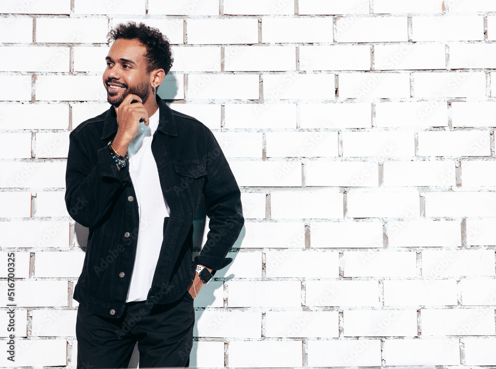 Handsome smiling hipster model. Sexy unshaven man dressed in black summer shirt and jeans clothes. Fashion male posing near white brick wall in studio at sunny day. Isolated Handsome smiling hipster model. Sexy unshaven man dressed in black summer shirt and jeans clothes. Fashion male posing near white brick wall in studio at sunny day. Isolated