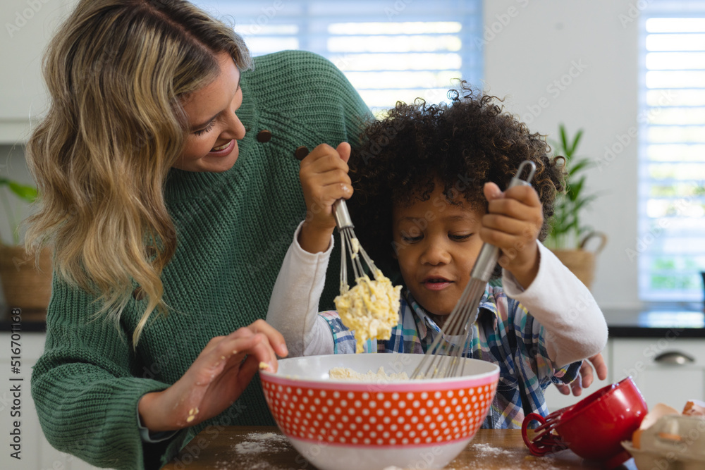 Happy multiracial mother looking at son with afro mixing batter in bowl with wire whisks in kitchen