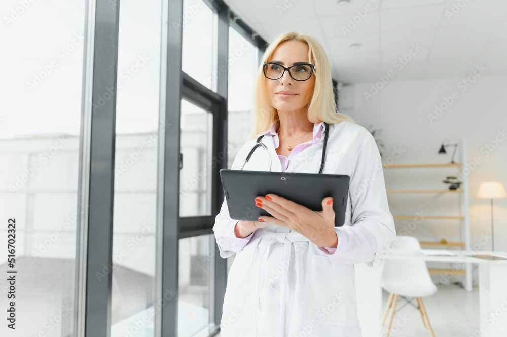 Portrait of middle age female doctor is wearing a white doctor's coat with a stethoscope around her neck. Smiling physician standing at private clinic