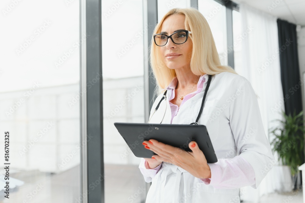 Portrait of beautiful mature woman doctor holding digital tablet. Confident female doctor using digital tablet with colleague talking in background at hospital. Portrait of beautiful mature woman doctor holding digital tablet. Confident female doctor using digital tablet with colleague talking in background at hospital.