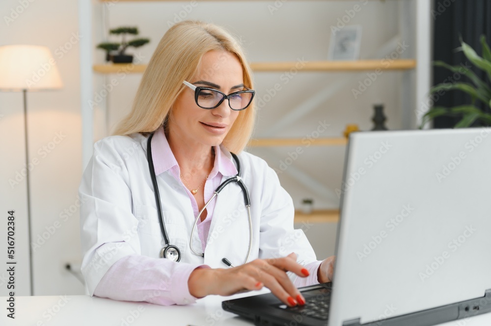 Portrait of middle age female doctor is wearing a white doctor's coat with a stethoscope around her neck. Smiling physician standing at private clinic Portrait of middle age female doctor is wearing a white doctor's coat with a stethoscope around her neck. Smiling physician standing at private clinic