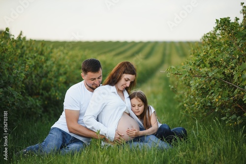 Beautiful pregnant brunette mom and dad and dauther in white clothes look at each other in nature.