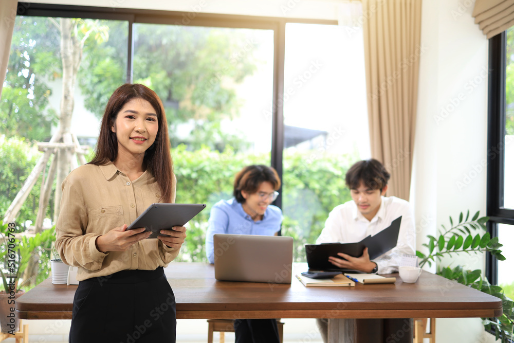 Portrait of a woman holding a tapet in the office with a coworker in the background.