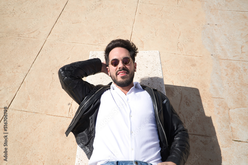 Obraz premium Portrait of handsome young man with beard, sunglasses, leather jacket and white shirt, lying on a stone bench. Concept beauty, fashion, trend, peace, relax, tranquility.