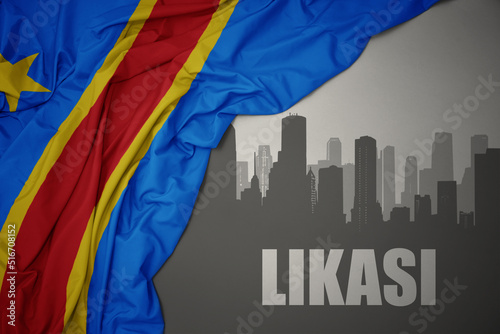 abstract silhouette of the city with text Likasi near waving colorful national flag of democratic republic of the congo on a gray background. photo