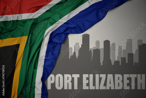 abstract silhouette of the city with text Port Elizabeth near waving colorful national flag of south africa on a gray background.