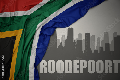 abstract silhouette of the city with text Roodepoort near waving colorful national flag of south africa on a gray background. photo