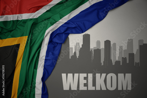 abstract silhouette of the city with text Welkom near waving colorful national flag of south africa on a gray background. photo