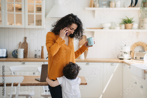 Картината върху платно Cute little boy embraces his mom  at kitchen while she is talking by phone holding cup, smiling, looking at son