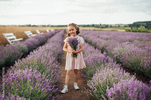 pretty smiling girl in pink dress is standing with a bouquet of flowers in hands among lavender filed