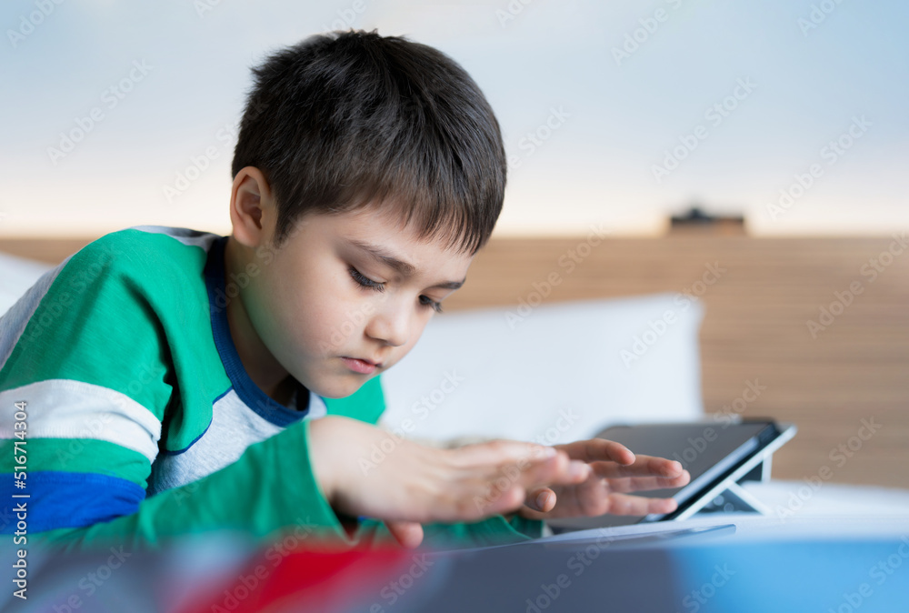 Cinematic portrait Kid playing games online on internet with friends in bed room, Young boy using tablet typing or chatting at night, Childhood sitting on bed relaxing at home on weekend. Cinematic portrait Kid playing games online on internet with friends in bed room, Young boy using tablet typing or chatting at night, Childhood sitting on bed relaxing at home on weekend.