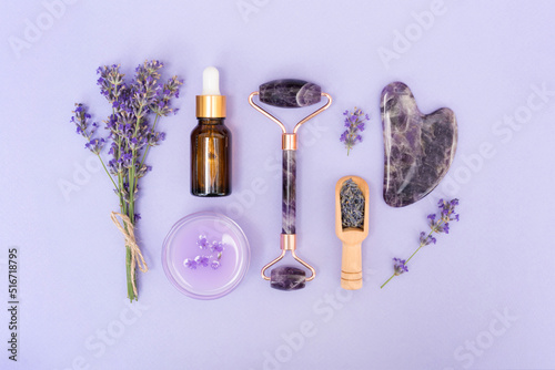 Amethyst gua sha roller and scraper for facial massage with bottle of face serum or lavender cosmetic oil and moisturizer over purple background. Facial treatments kit and skincare concept