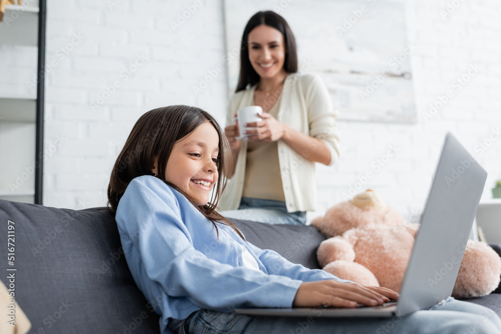 excited girl gaming on laptop near nanny with tea cup on blurred background. excited girl gaming on laptop near nanny with tea cup on blurred background.