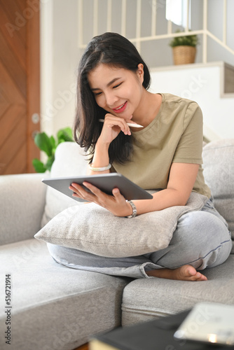 Smiling young woman reading ebook on digital tablet while relaxing in comfortable couch at home