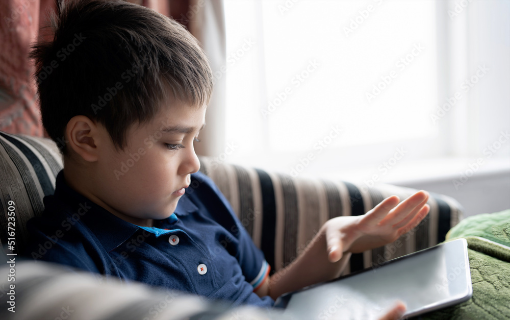Cinematic portrait young boy playing game on tablet sitting on sofa with light shining from window, Kid playing games online on internet at home, Child talking video call with friends at home Cinematic portrait young boy playing game on tablet sitting on sofa with light shining from window, Kid playing games online on internet at home, Child talking video call with friends at home