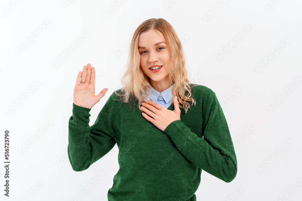 Lovely fair-haired young woman raising hand and holding palm on heart, making promise, looking seriously, wears casual clothes. Indoor studio shot on white background. Lifestyle concept