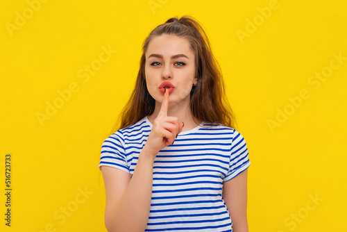 Attractive brunette girl holding index finger at her mouth, making shush sign, saying Shh, dont tell anyone, asking you to keep her secret, stands in casual striped t shirt over yellow background