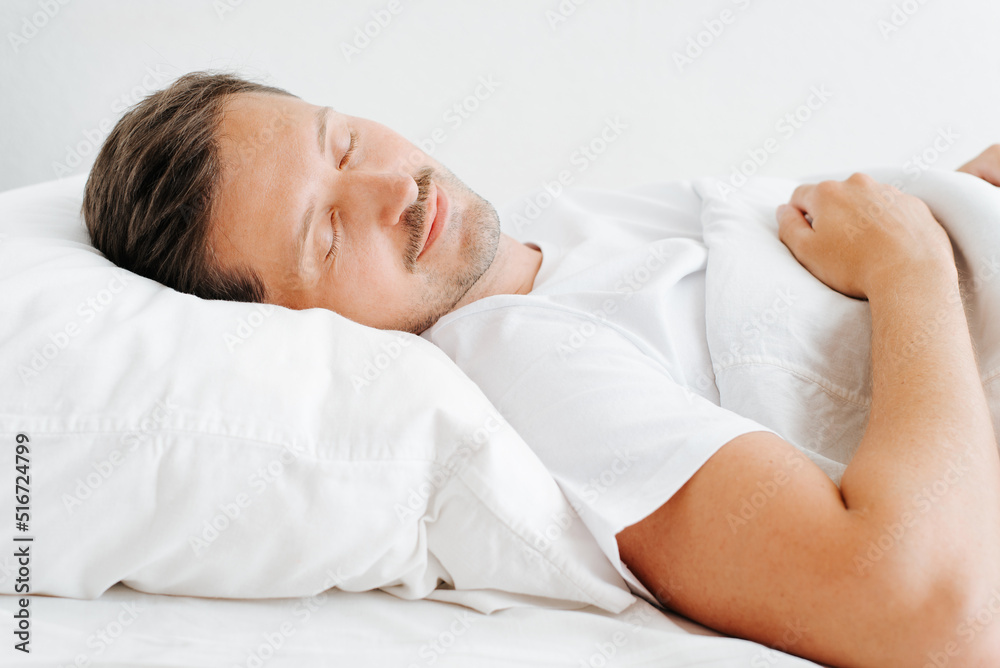 Smiling sleeping man in bed, side view. Resting guy with closed eyes lying in bedroom indoors in early morning Smiling sleeping man in bed, side view. Resting guy with closed eyes lying in bedroom indoors in early morning