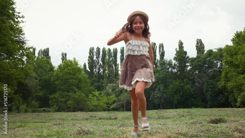 Happy Caucasian little girl in a straw hat and dress running skipping through the lawn in the park. Summer holidays. Slow motion. The concept of childhood and Children's Day.