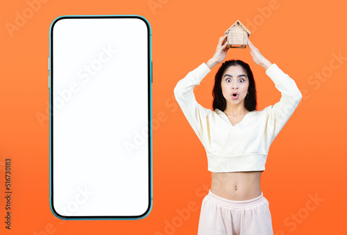 Big blank smartphone with white mockup screen and shocked indian race woman holding a little toy house above her head. Dream of a house. Own home concept. Real estate advertisement background. photo