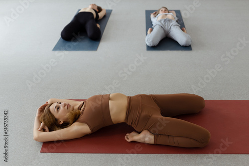 Slim young woman lying in Supta Virasana, reclining Hero Pose, stretching abdominal and back muscles on mat in gym photo