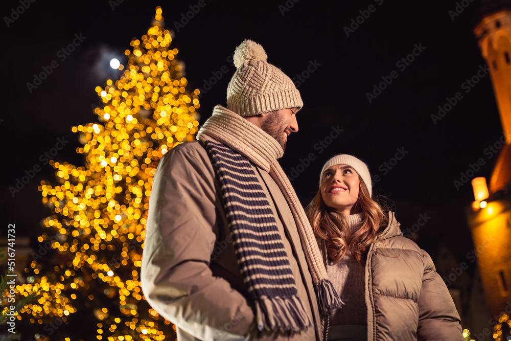 Naklejka premium winter holidays and people concept - happy smiling couple over christmas tree lights in evening city