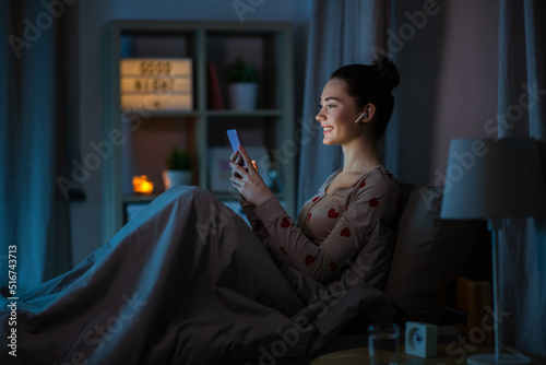 technology, bedtime and people concept - happy smiling teenage girl with smartphone and earphones sitting in bed at home at night