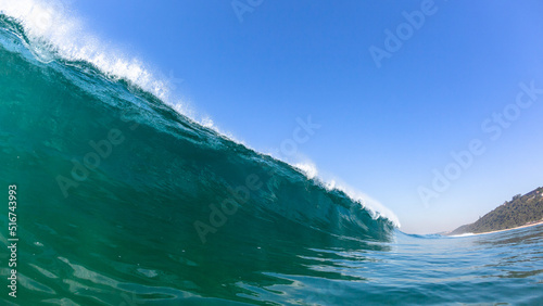 Ocean wave swimming close up encounter perspective of long sea wall of water about to crash break towards beach coastline with blue clear sky.