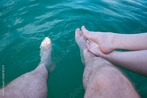 Fototapeta Naklejka Na Ścianę i Meble -  Unrecognizable caucasian woman and man dipping their bare feet in clean and clear lake water. Sunny summer day