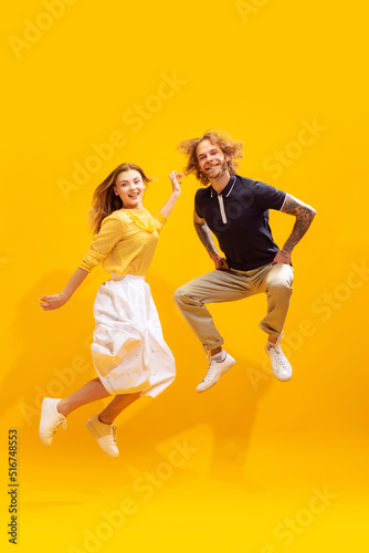Portrait of young man and woman in casual clothes posing, cheerfully jumping isolated over yellow studio background