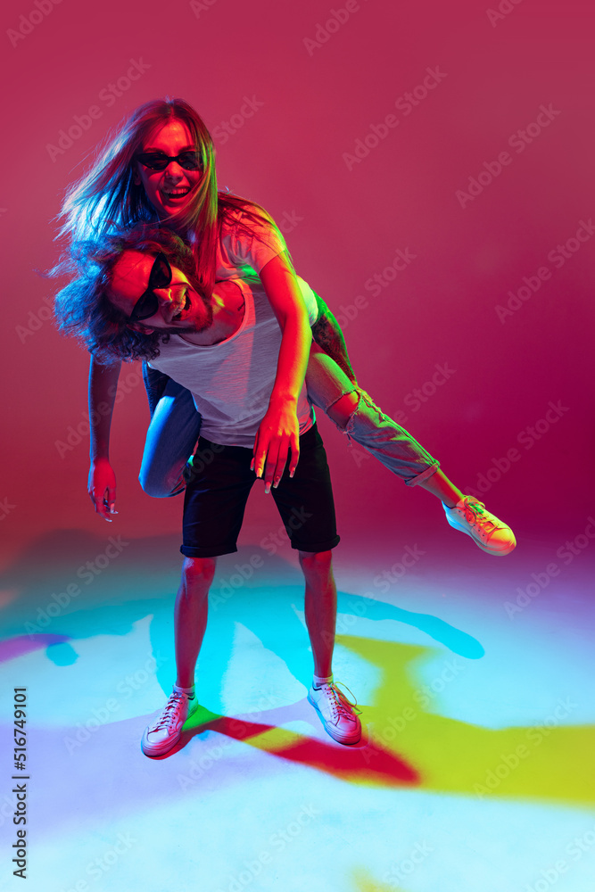 Obraz premium Portrait of young stylish man and woman having fun, posing isolated over red background in neon light