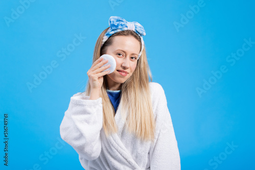 Cleansing, washing morning face care towel-wiping delicate skin. Portrait of young women in a bathrobe on a blue studio background. © ABCreative
