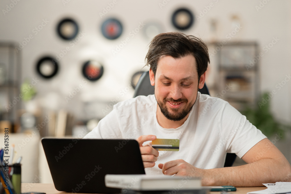 Cheerful young guy orders gift to his girlfriend. He prefers to shop online because it saves time. In front of him is laptop. He holds bank card and places an order. Cheerful young guy orders gift to his girlfriend. He prefers to shop online because it saves time. In front of him is laptop. He holds bank card and places an order.