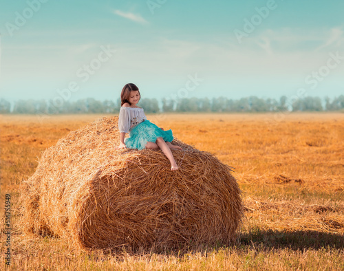 The girl in sits on a roll, stack of hay. The girl is walking in the field, it is a hot summer. The girl is wearing a blue skirt.