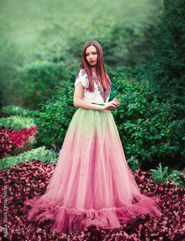 A slender girl stands among pink flowers in a long puffy dress.
Around the lush foliage, many flowers, fabulous forest. The girl has long hair and makeup.