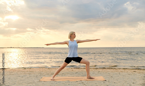 fitness, sport, and healthy lifestyle concept - happy woman doing yoga warrior pose on beach over sunset