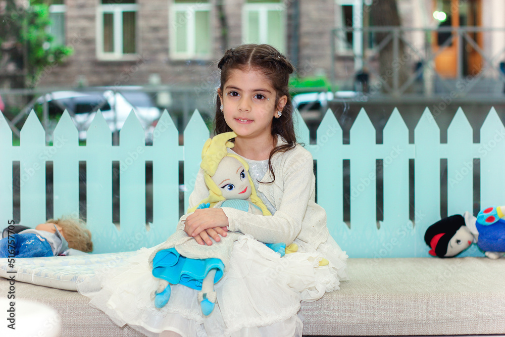 Adorable girl hugging her handmade soft toy and sitting on the floor in kindergarten with multi colored interior