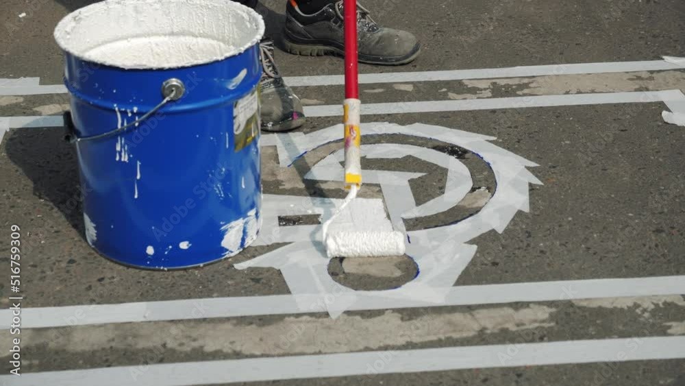 Close-up of manual utility male worker dipping roller brush in paint metal bucket, painting white marking line of disabled car parking lot sign on asphalt, drawing on road surface, summer daytime. Close-up of manual utility male worker dipping roller brush in paint metal bucket, painting white marking line of disabled car parking lot sign on asphalt, drawing on road surface, summer daytime.