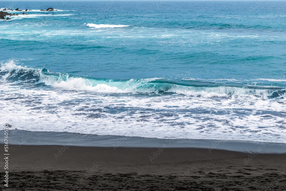 Waves with white foam on the crests of the sea with turquoise blue water and black sand on the beach. Coastline of Atlantic Ocean in the Canary Islands, Spain. Summer resort background with copy space