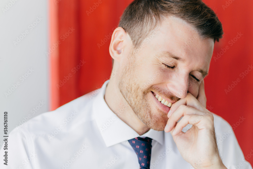 Joyful unshaven male with happy expression, giggles at funny joke, closes eyes with pleasure, dressed in elegant white shirt, recieves pleasant news, poses against red wall. Positive emotions concept Joyful unshaven male with happy expression, giggles at funny joke, closes eyes with pleasure, dressed in elegant white shirt, recieves pleasant news, poses against red wall. Positive emotions concept