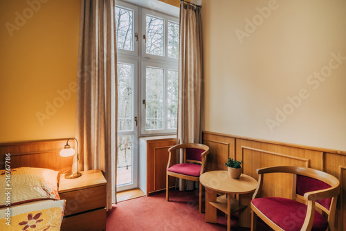 Old hotel room with two individual beds pushed together. Room has standard equipment in form of lamps, bedside tables and a seating area. Room also has window and yellow-painted walls with wood trim. © Zdena Venclik