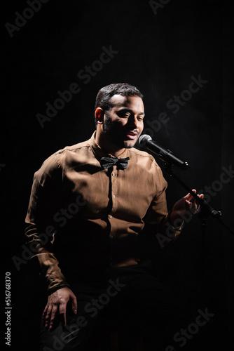 indian stand up comedian telling jokes into microphone on dark stage isolated on black.