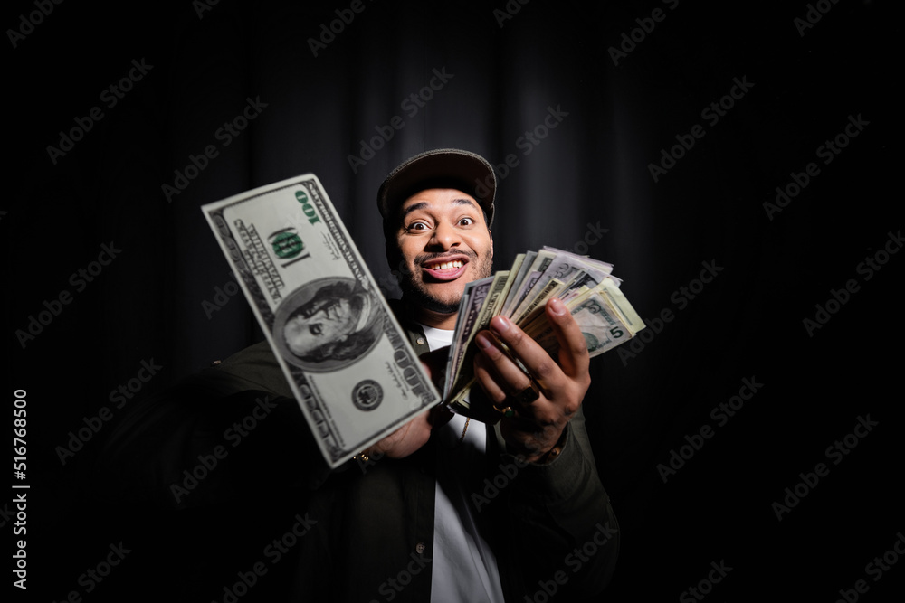 rich indian hip hop performer in cap throwing dollar banknotes and looking at camera on black. - obrazy, fototapety, plakaty 