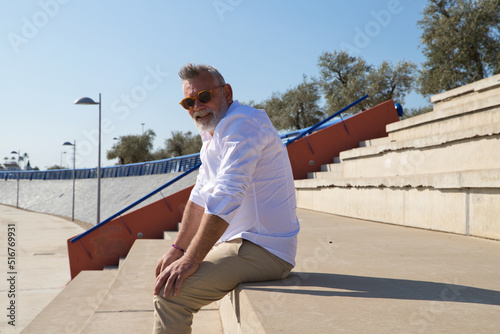 handsome mature man with beard and grey hair is sitting on the steps of an open-air auditorium. The man looks at different places. Senior concept, travel and tourism. © @skuder_photographer