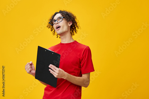 Joyful happy myopic young student man in red t-shirt funny eyewear holds tablet folder with study notes posing isolated on over yellow studio background. Free place for ad. Education College concept