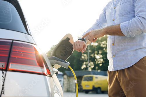 Unrecognizable businessman plugging in charging cable to to electric vehicle. Male hand inserts power connector into EV car and charges batteries, uses smartwatch for activates start charging.